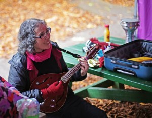 A white woman with glasses and curly hair plays mandolin in a park