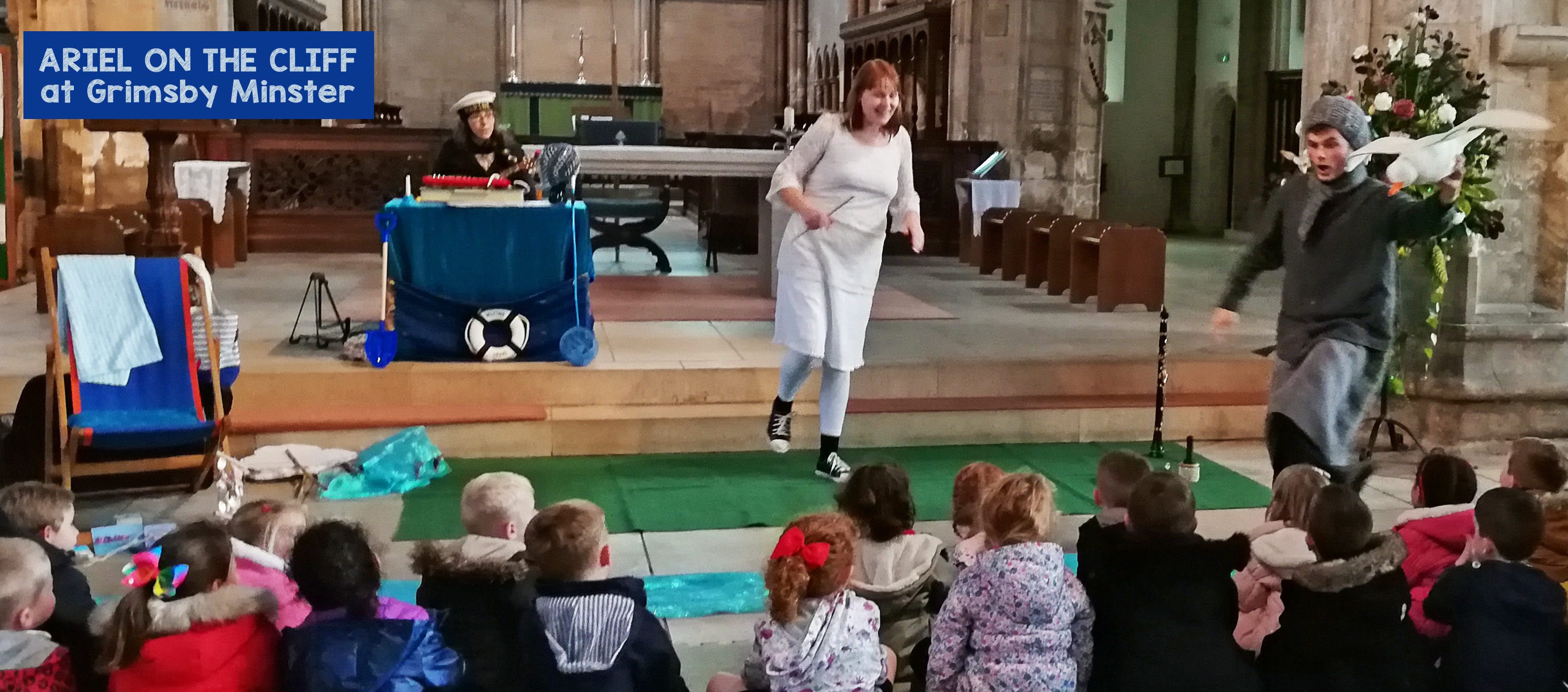 Three white performers with a sea themed set play instruments and puppet a seagull
