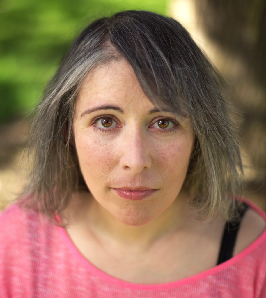 A white woman with straight greying shoulder length hair and a pink jumper